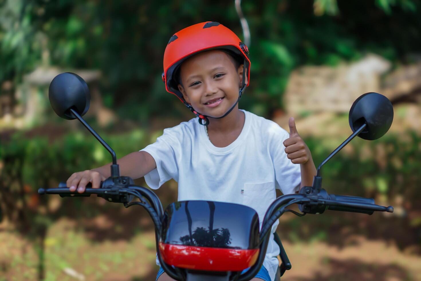 A young asian boy in a red helmet gives a thumbs up while sitting on a electric bike in outdoor photo