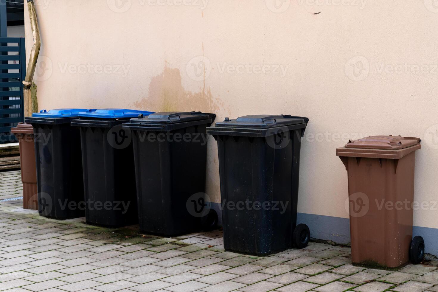 Various trash bins in different colors are lined up against a building photo