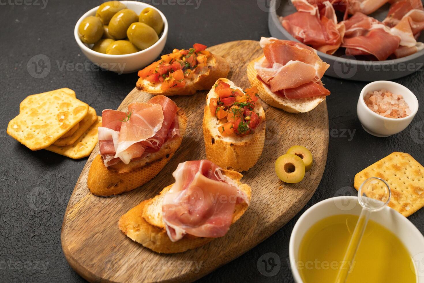 Italian sandwiches bruschetta with olives, bran bread, cherry tomatoes, prosciutto on wooden cutting board, dark black background surface photo