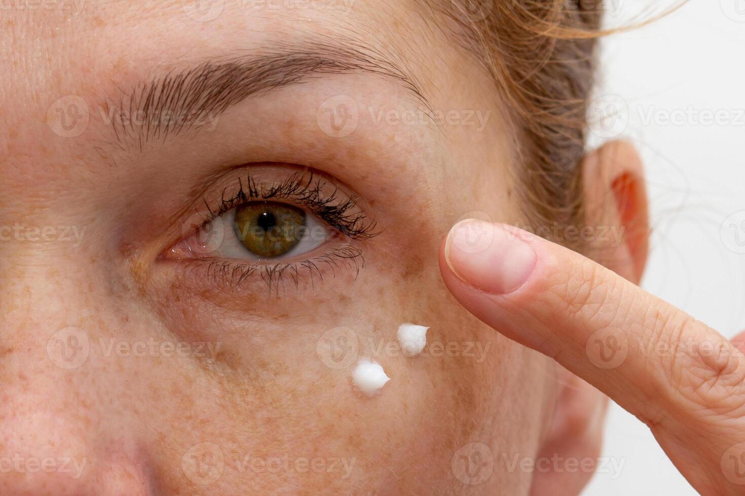 Close up cropped eye of middle aged caucasian woman looking at camera applying cream on skin photo