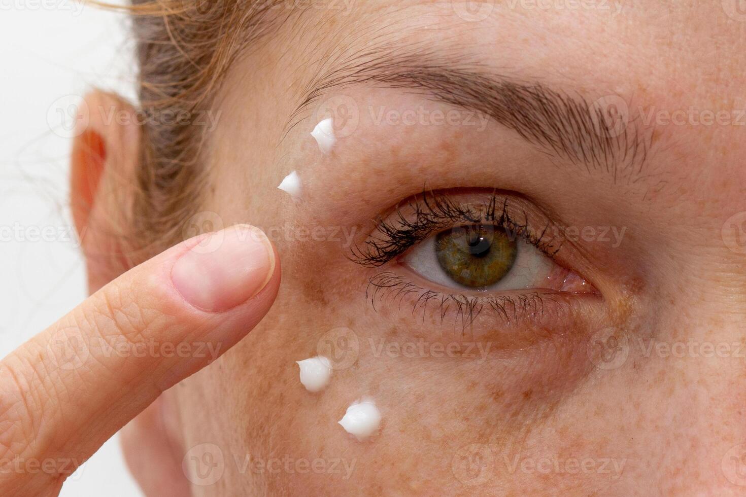 Close up cropped eye of middle aged caucasian woman looking at camera applying cream on skin photo