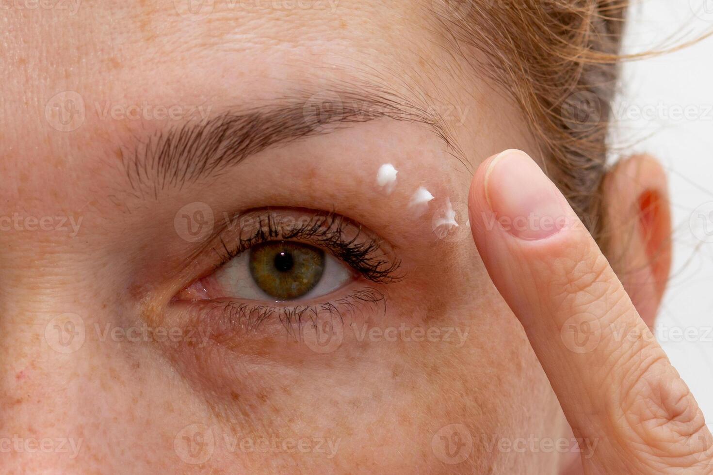 Close up cropped eye of middle aged caucasian woman looking at camera applying cream on skin photo