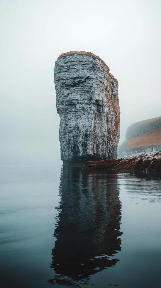 Mysterious rock formation rises from calm water on foggy morning near coastal cliffs photo