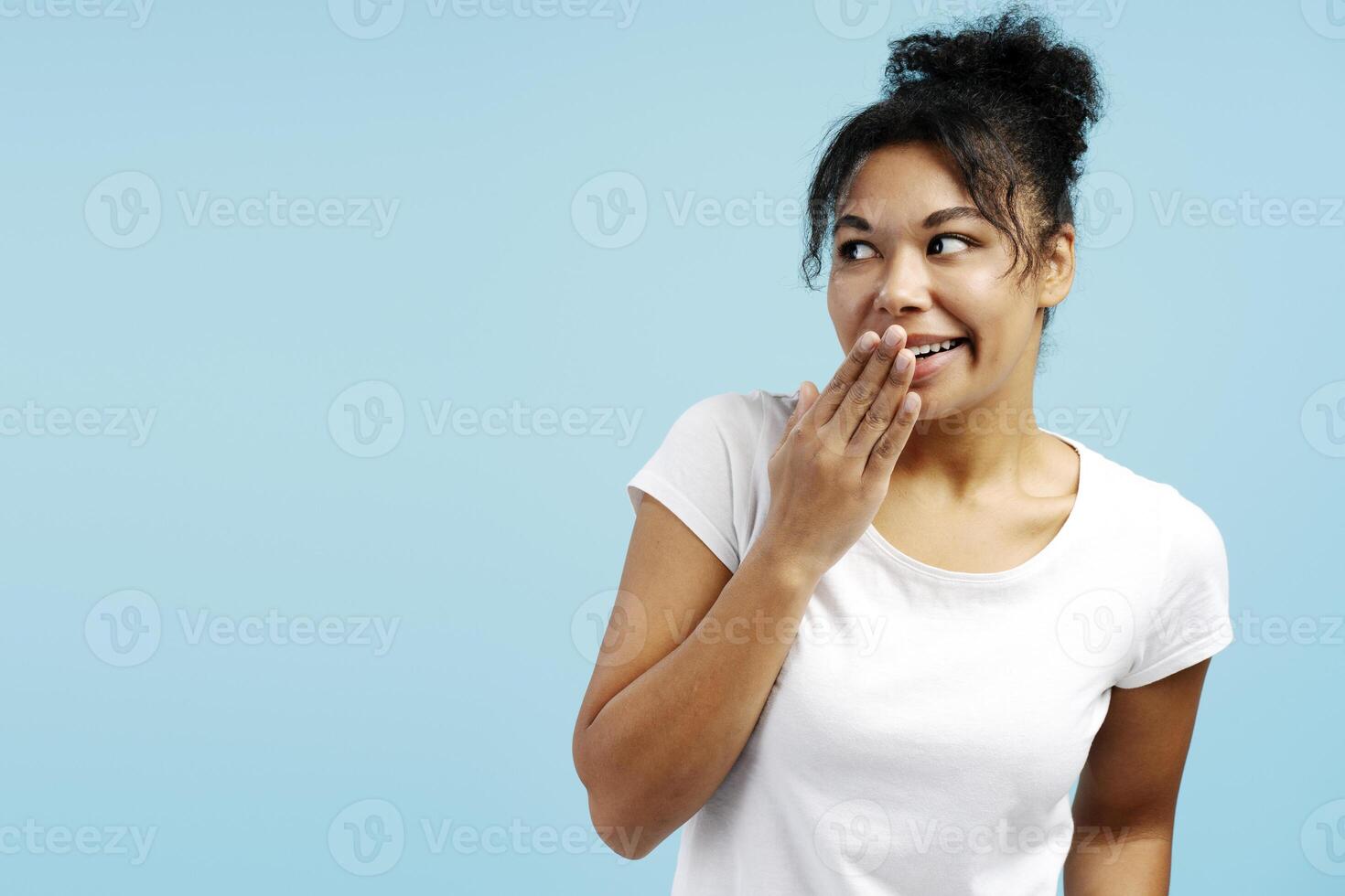 Young woman covering mouth with hand and looking away on light blue background photo