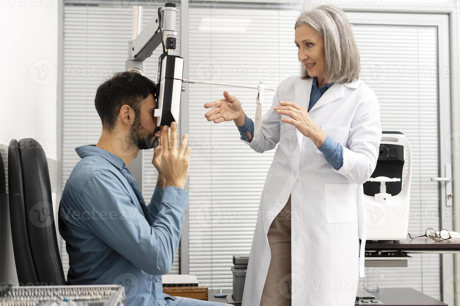 Senior female doctor performing eye exam on male patient using phoropter in clinic photo