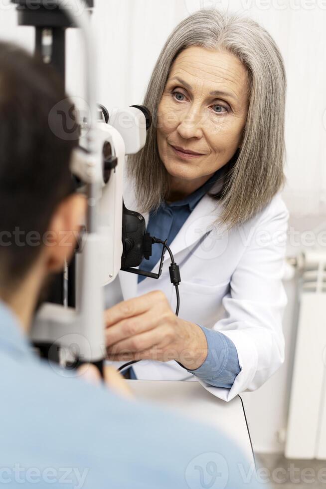 Senior female doctor performing eye exam on patient using phoropter in ophthalmology clinic photo