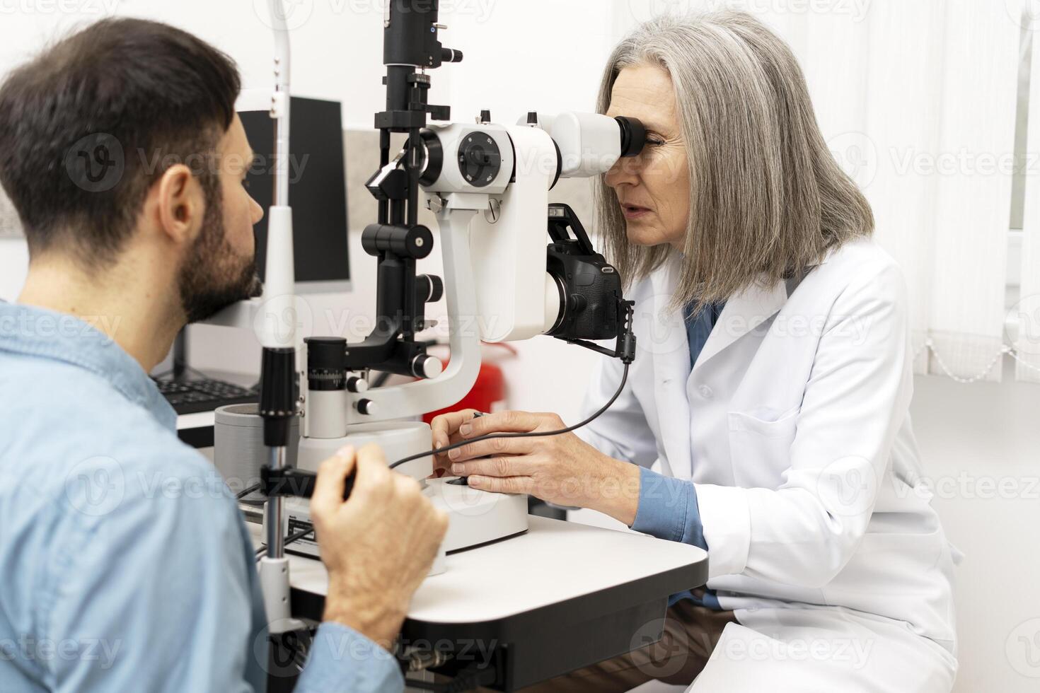 Senior female doctor performing eye exam on male patient using slit lamp photo