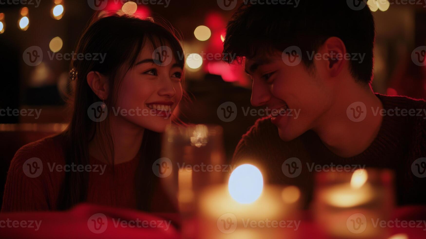 A couple shares a joyful moment during a candlelit dinner, surrounded by warm lighting and vibrant decorations. Their smiles indicate a deep connection as they gaze into each other's eyes. photo