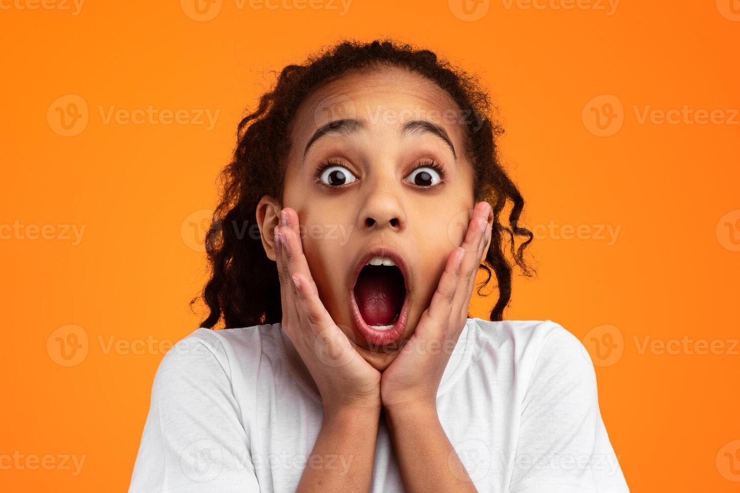 Wow. Closeup portrait of excited African American girl touching her cheeks, looking and staring at camera with open mouth, surprised by sale, isolated over yellow studio background photo