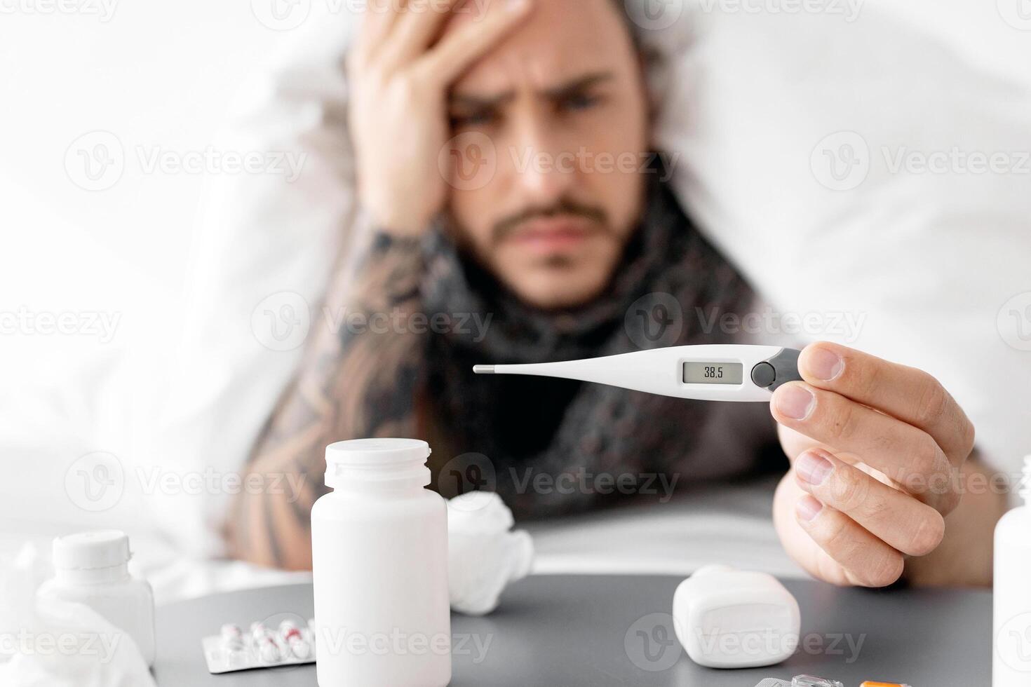 Coronavirus and influenza symptoms. Sick guy showing thermometer with high temperature result to camera, lying on bed with table full of pills nearby photo