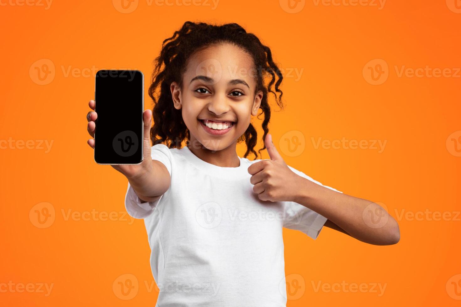 Great Mobile App. Portrait Of Positive Cheerful African American Girl Holding And Showing Smartphone Empty Screen Gesturing Thumbs Up Like Over Yellow Background. Bright Studio Shot, Mockup photo