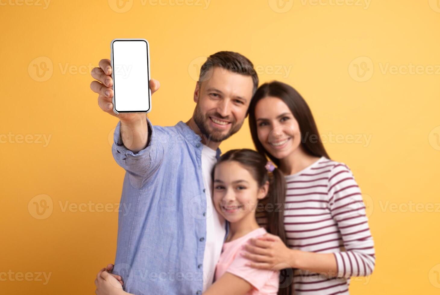A father and mother embrace their teenage daughter, smiling joyfully in a vibrant yellow backdrop. They share a moment of love, capturing a family memory with a smartphone. photo