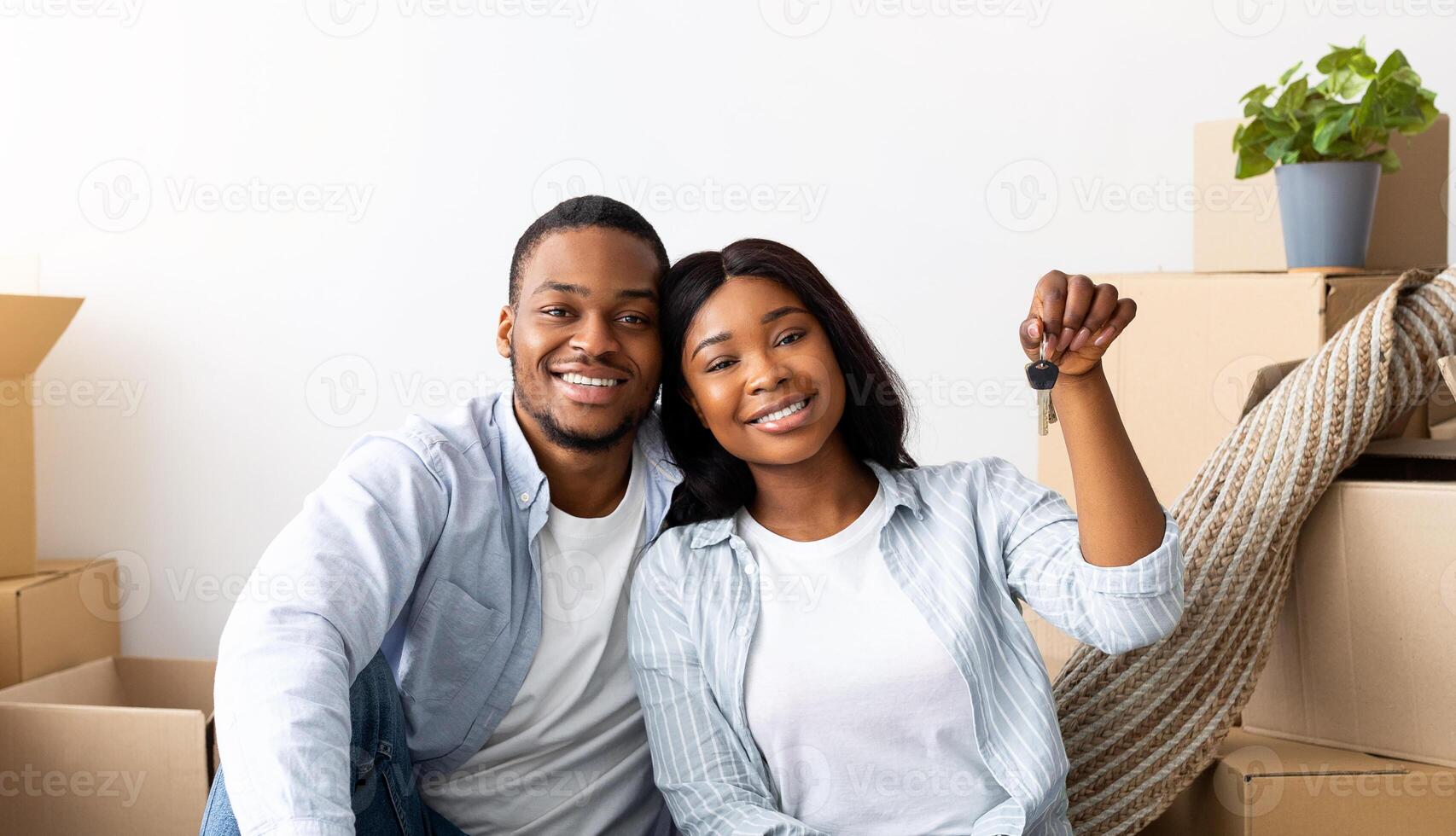 Happy african american spouses with house key and cardboard boxes looking at camera and smiling, sitting on floor in their property. Young black homeowners being happy to move into own apartment photo