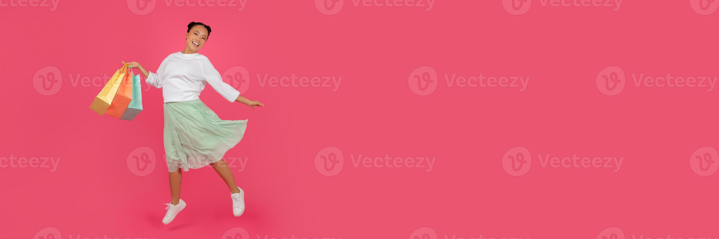 Shopping Day Concept. Full length of happy asian lady jumping with colorful paper bags, positive korean lady carrying new purchases, having fun on pink studio background, full length, copy space photo