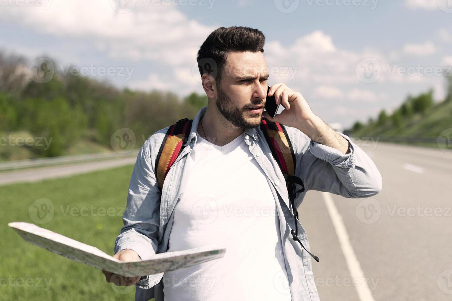 Handsome young man standing on road with map, feeling lost, calling on smartphone, checking travel options, empty space. Stressed millennial guy hitchhiking on highway, cannot catch ride photo