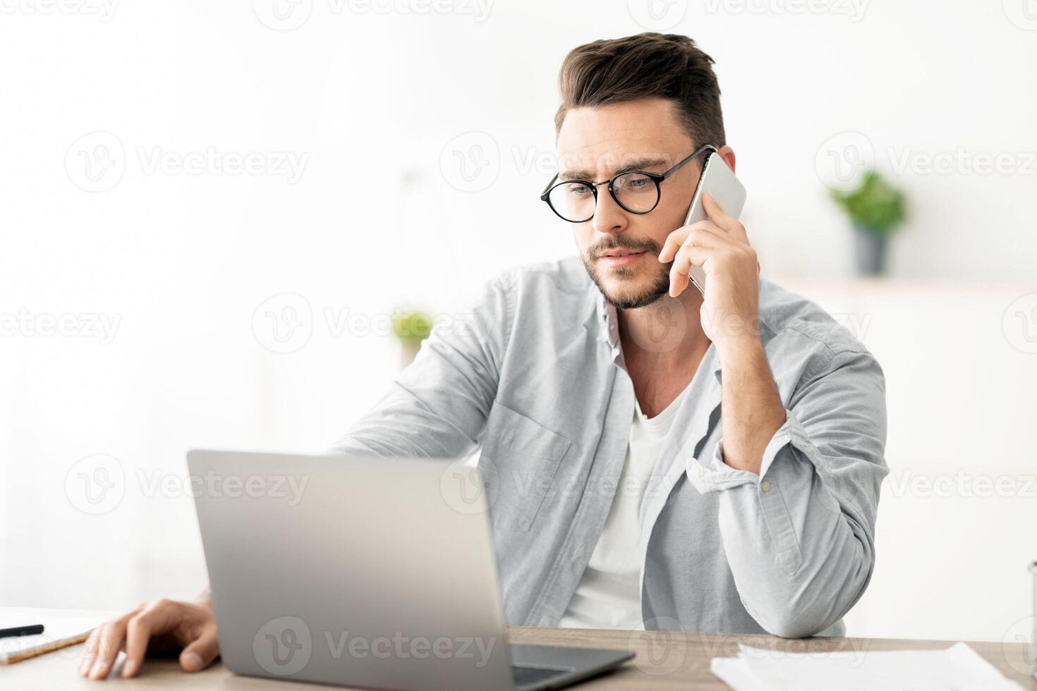 Handsome young man talking on smartphone, speaking with customer, working on laptop at home office workplace. Entrepreneur having business call conversation. Communication Concept photo