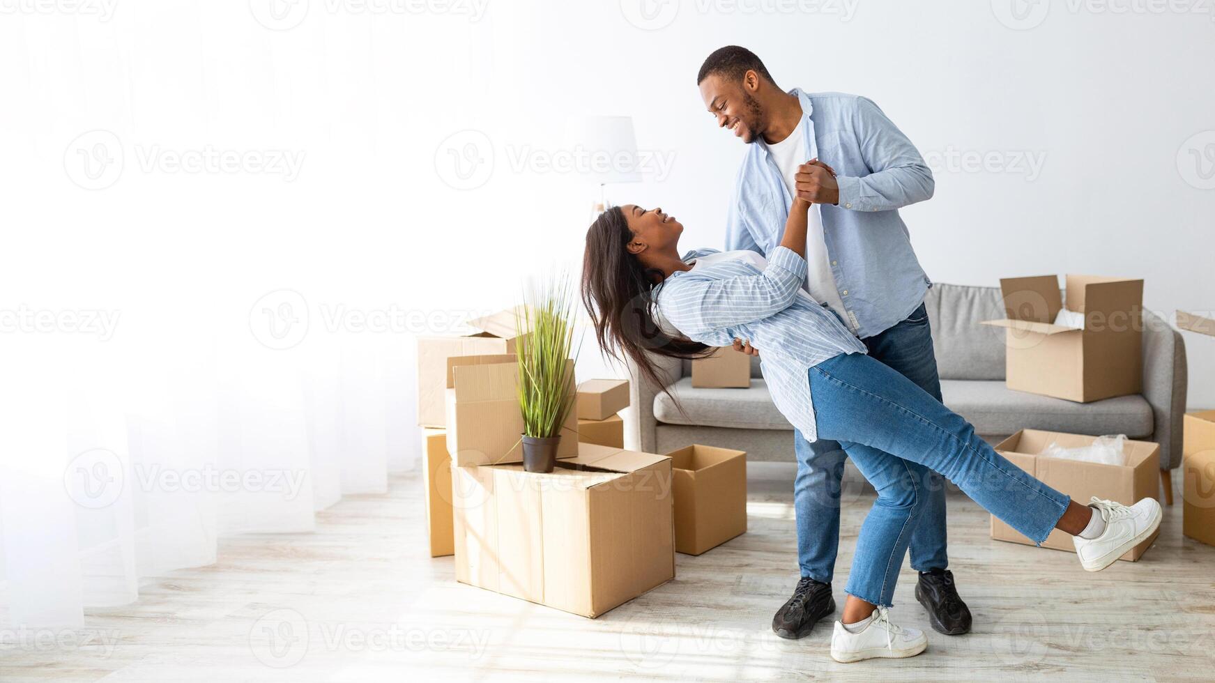 Happy loving african american couple dancing in their new house among carton boxes on moving day, panorama. Excited young family having fun while relocating to new property photo