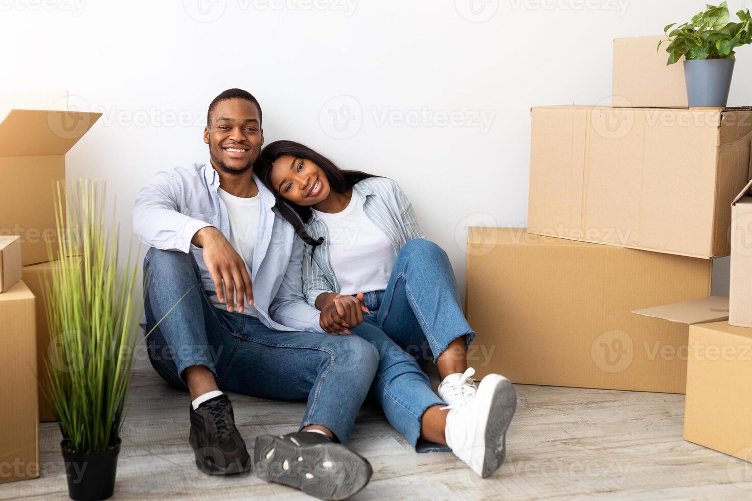 Real estate rent concept. Loving african american spouses relocating to new apartment, sitting among cardboard boxes at new home and smiling to camera, free space. Mortgage and ownership photo