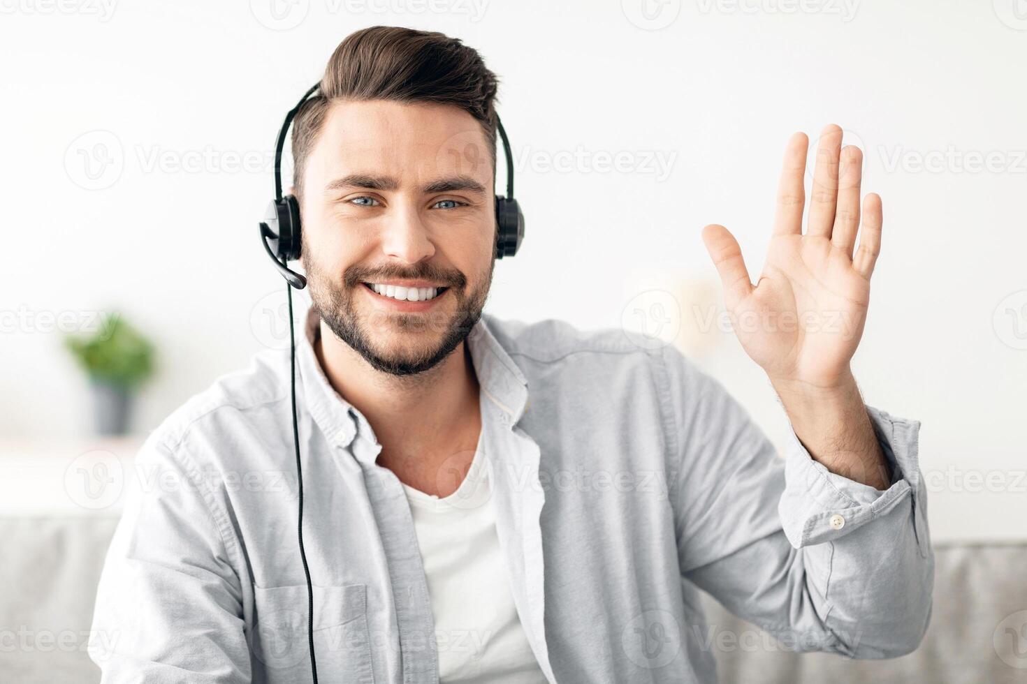 Online teacher's portrait. Happy handsome man wearing headset waving hand to webcam, gesturing hello and smiling while sitting on sofa at home. Modern distance tutoring and e-teaching concept photo