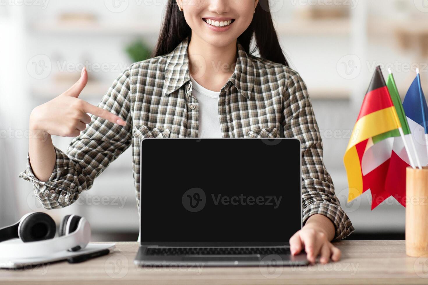 Happy asian woman pointing at laptop with blank screen, sitting at worktable with flags of different countries. Cheerful young lady recommending website for foreign languages learning, home interior photo