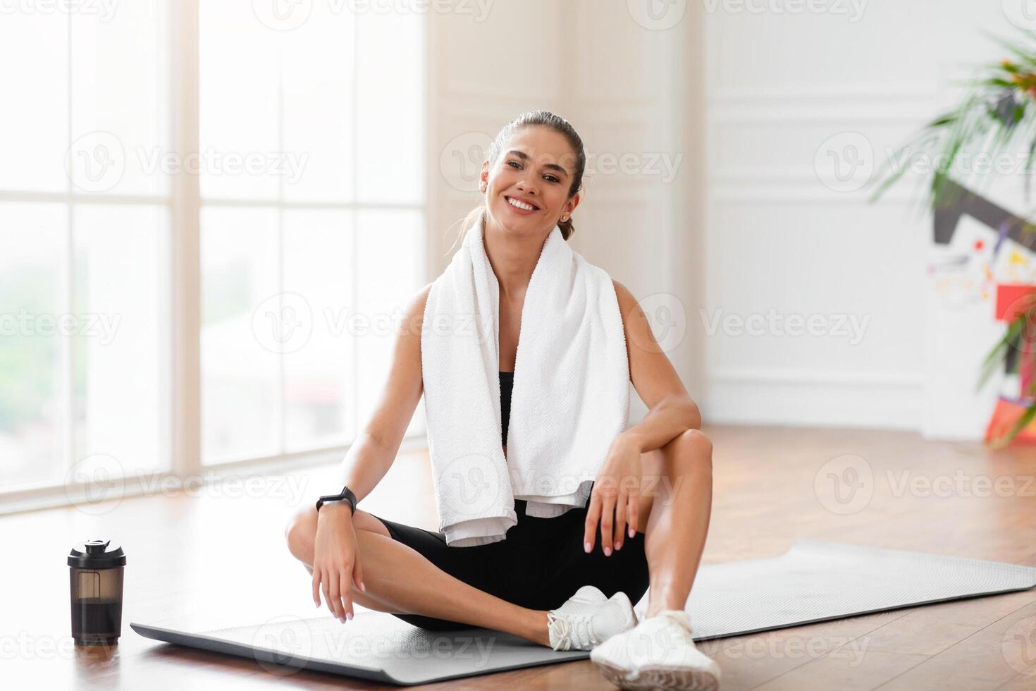 Portrait of sporty beautiful smiling lady in sportswear and white towel on neck sitting on the floor yoga mat near window and posing looking at camera, exercising in gym or at home in minimal interior photo