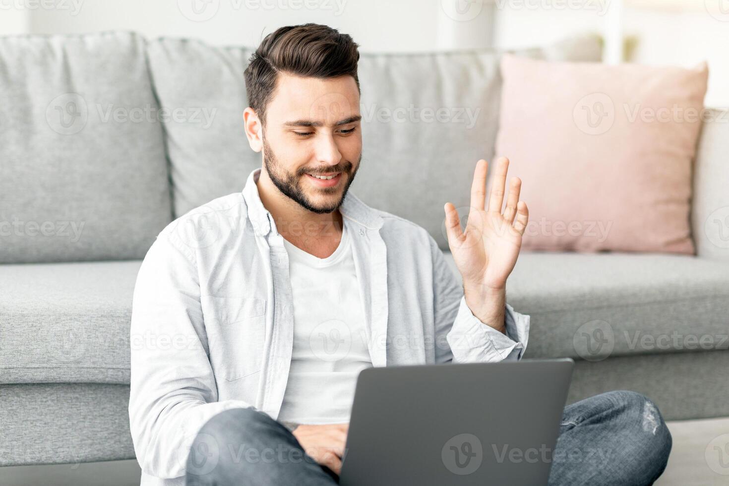 Happy millennial man waving hand to laptop calling while sitting on floor in living room interior, free space. Handsome guy communicating online using computer at home photo