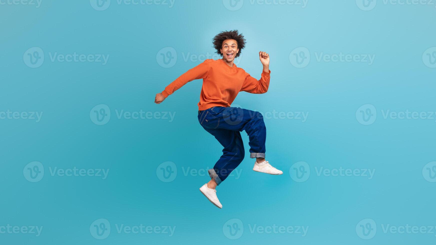 A young African American man with curly hair is jumping joyfully against a vibrant blue background. He is wearing a casual orange sweater and blue jeans, exuding youthful energy and enthusiasm. photo