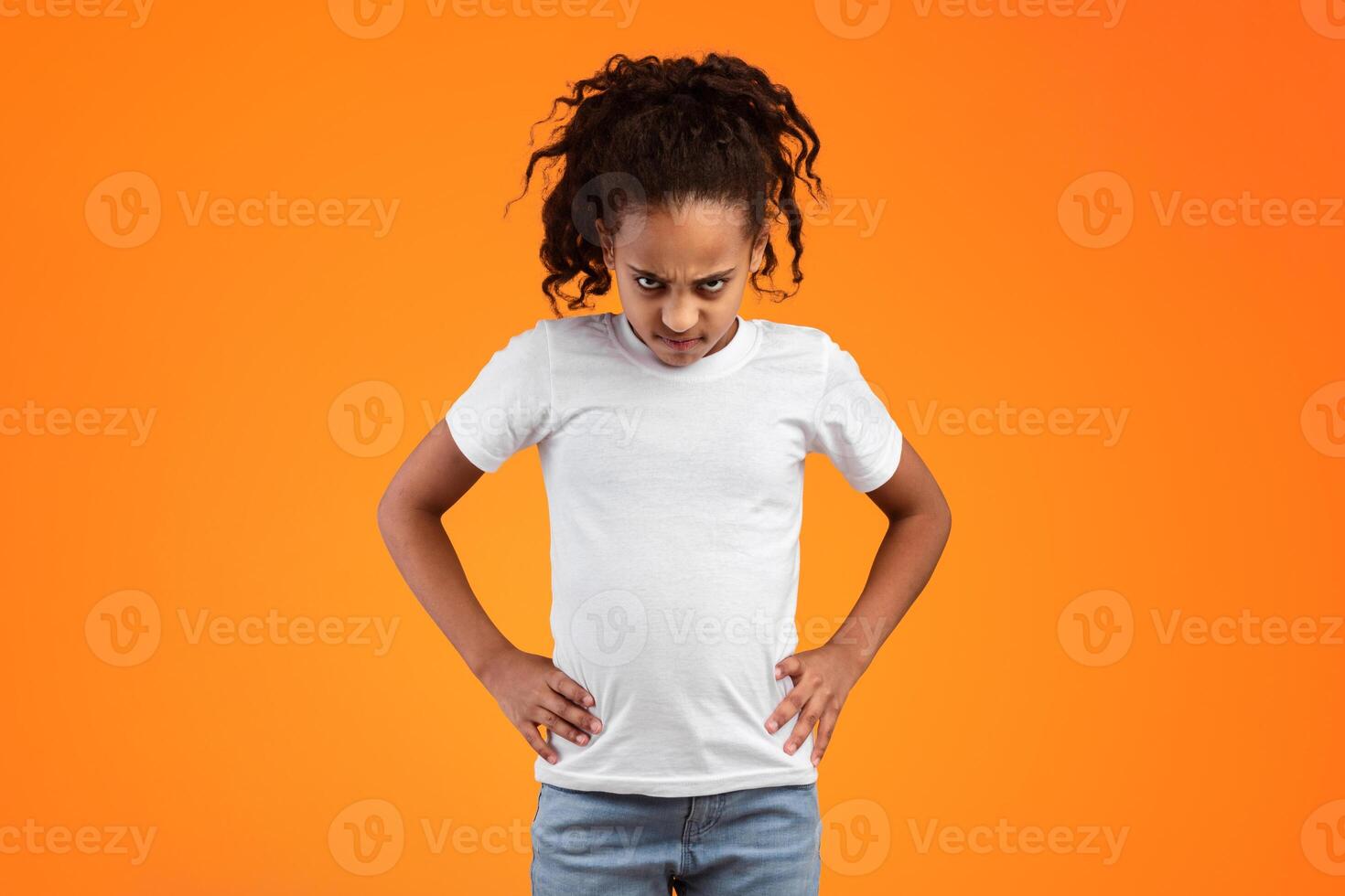 Grumpy Person. Serious furious black girl standing and putting hands on hips, posing over orange studio background, feeling angry and displeased, having bad mood, looking at camera and frowning photo