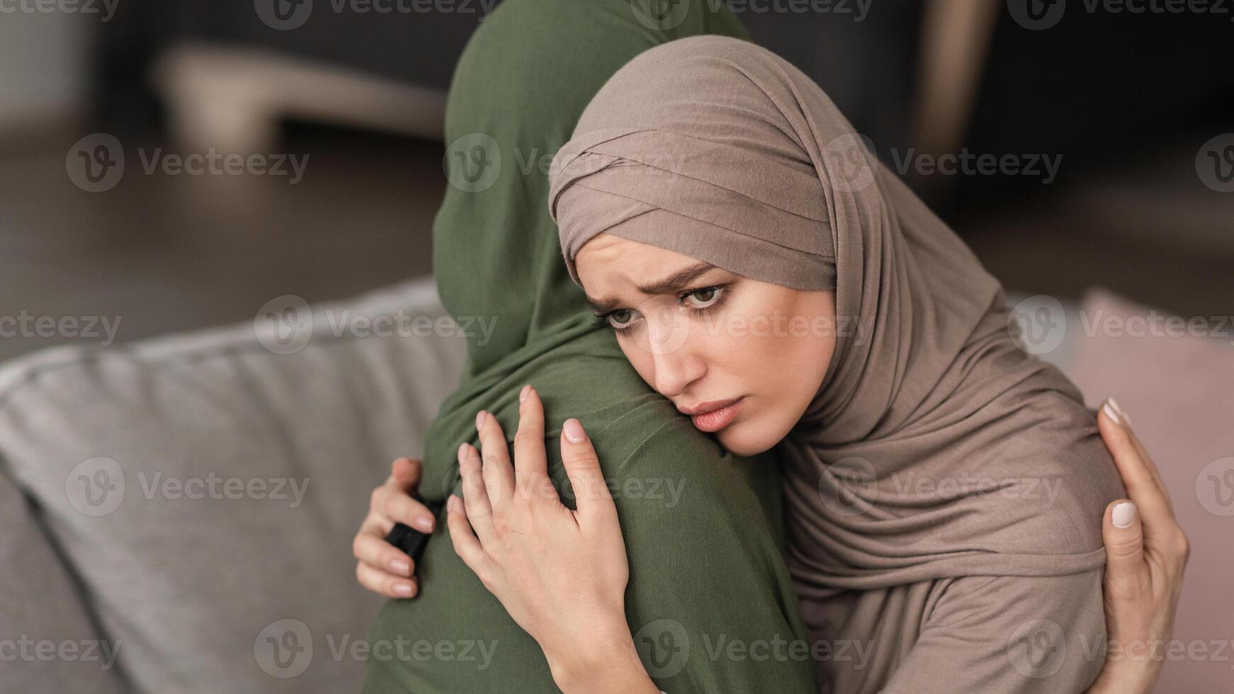 Unhappy Muslim Woman Hugging Depressed Senior Mother Supporting And Comforting Her Sitting On Sofa At Home, Wearing Hijab. Middle-Eastern Females Problems And Struggles Concept. Panorama photo