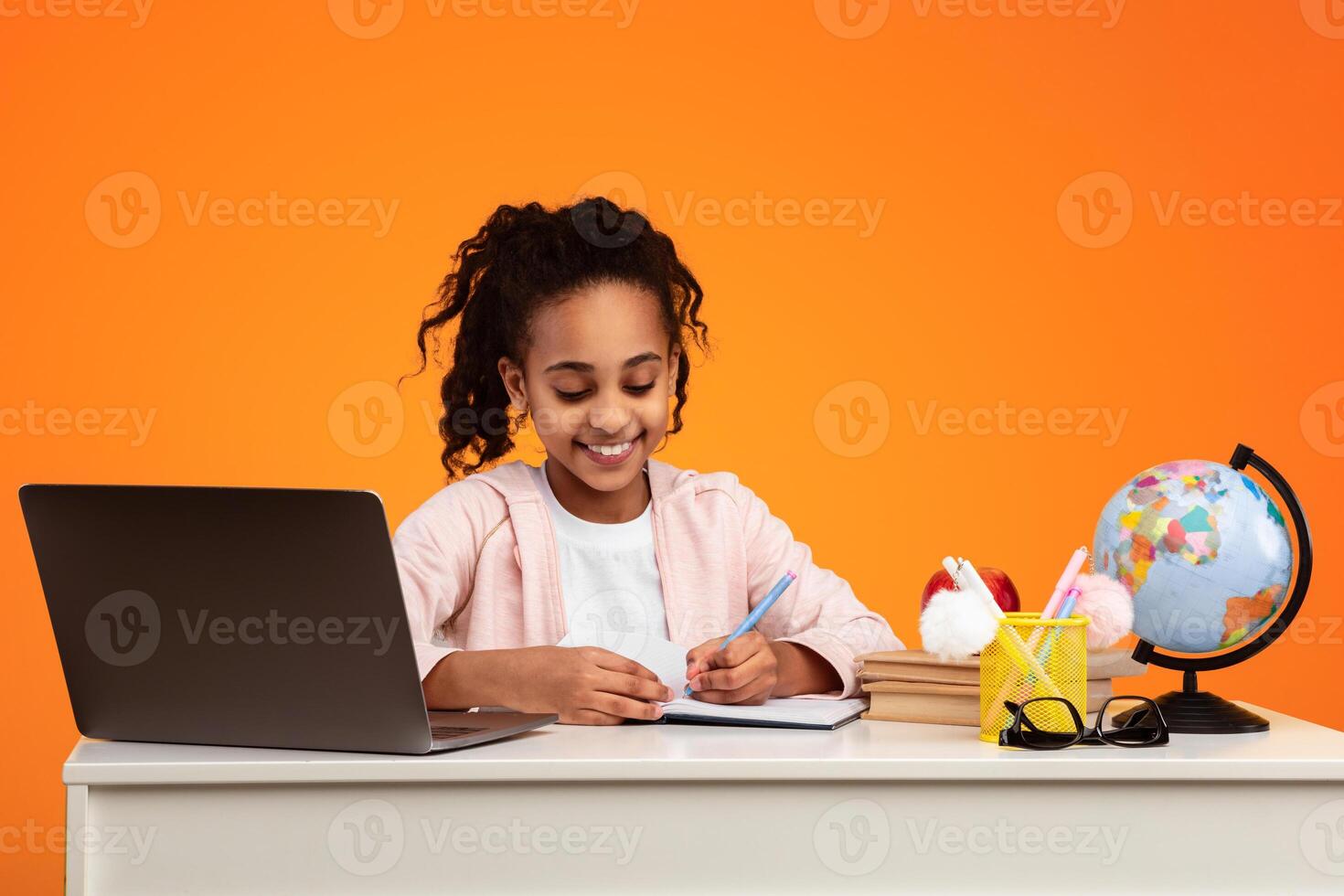 Modern Remote Education Concept. Portrait of smiling African American girl sitting at table, writing in notebook and using laptop computer. Positive young black student doing homework photo