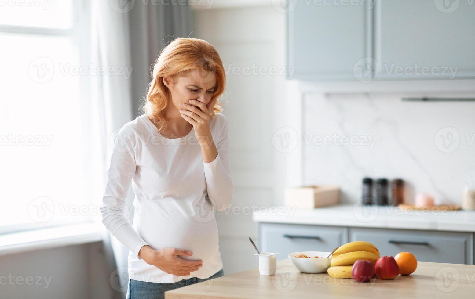 A pregnant woman stands in a modern kitchen, feeling nauseous while holding her stomach. She looks distressed as she glances at a bowl of fruits and a glass of yogurt on the countertop. photo