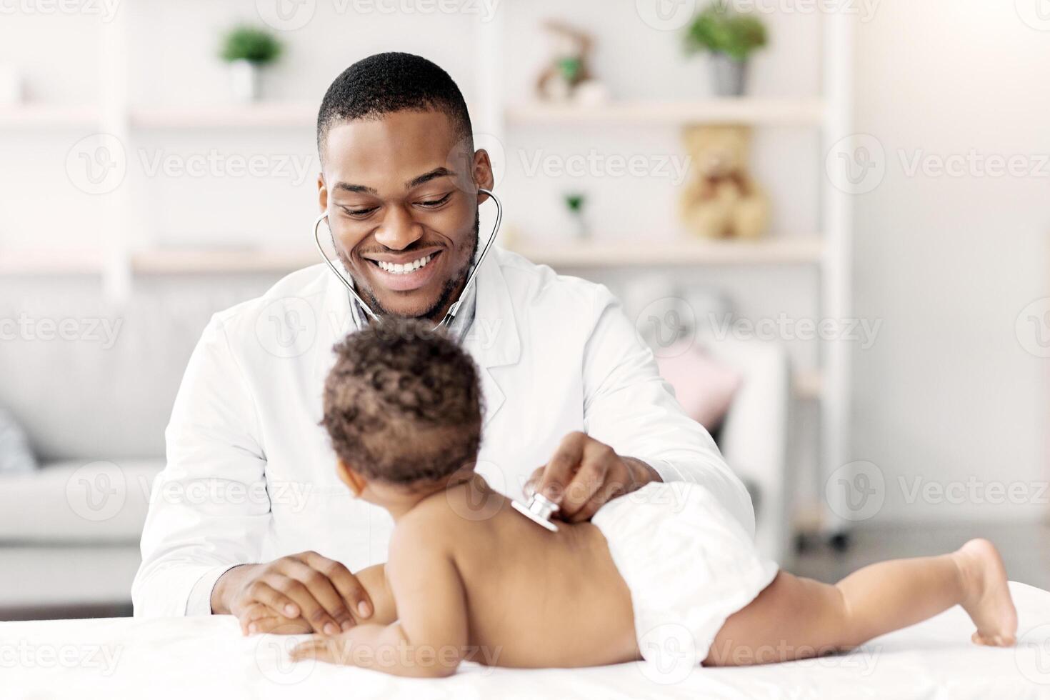 Medical Checkup. Smiling Black Pediatrician Doctor Examining Little Baby With Stethoscope During Appointment In Modern Clinic, Professional Therapist Checking Health Of Infant Child, Free Space photo