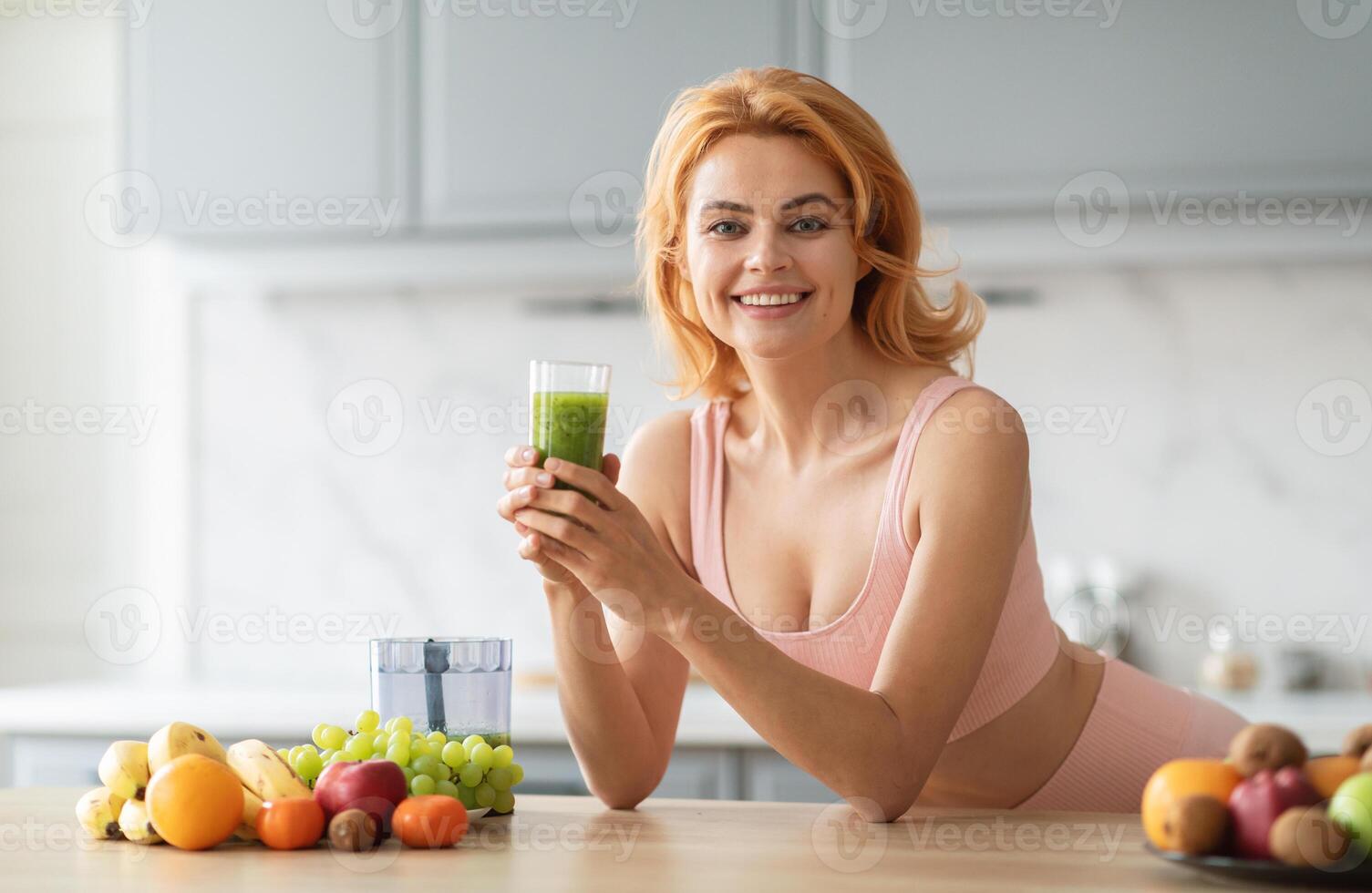 A young woman smiles while holding a glass of green smoothie in her kitchen. Fresh fruits are displayed on the countertop, creating a vibrant and healthy atmosphere. photo