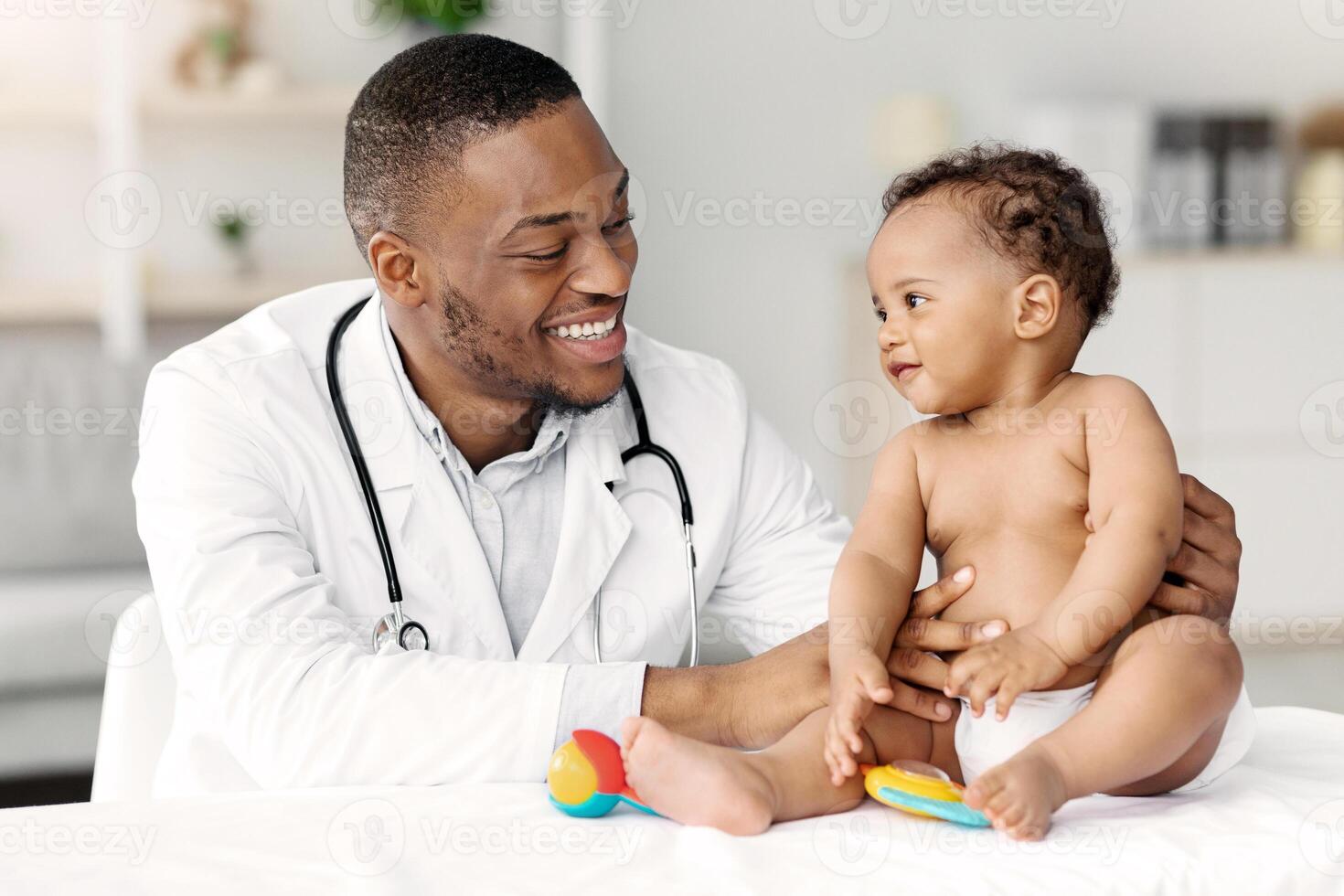 Child's Healthcare Concept. Portrait Of Smiling Black Doctor Making Check Up For Little Infant Baby Boy, Cute Toddler Child In Diaper Having Appointment At Pediatrician's Office, Free Space photo