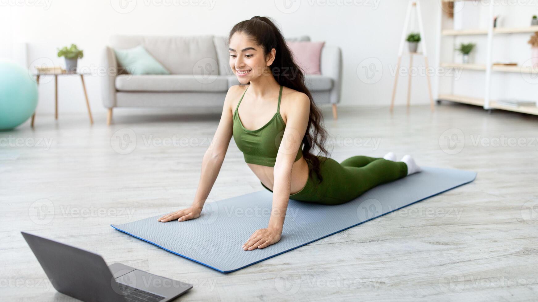 Full length of young Indian woman doing cobra asana, training to online yoga on laptop at home. Fit Eastern lady doing domestic workout for healthy spine during coronavirus lockdown photo