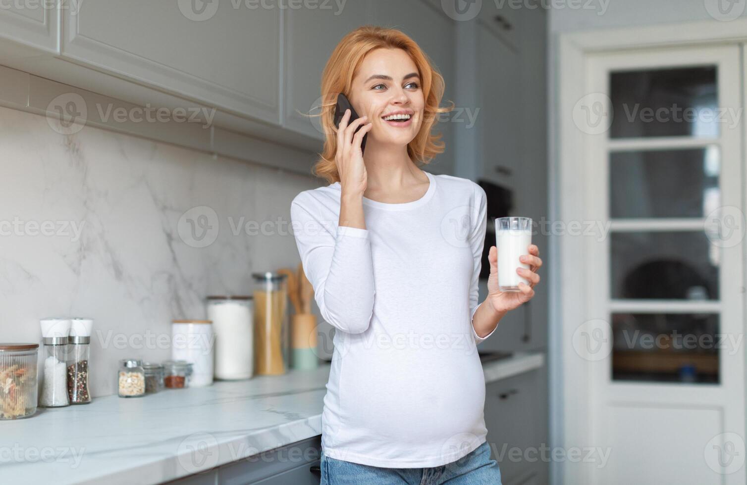 A cheerful pregnant woman stands in a well-lit kitchen, holding a glass of milk and smiling as she chats on the phone with someone, enjoying the moment. photo