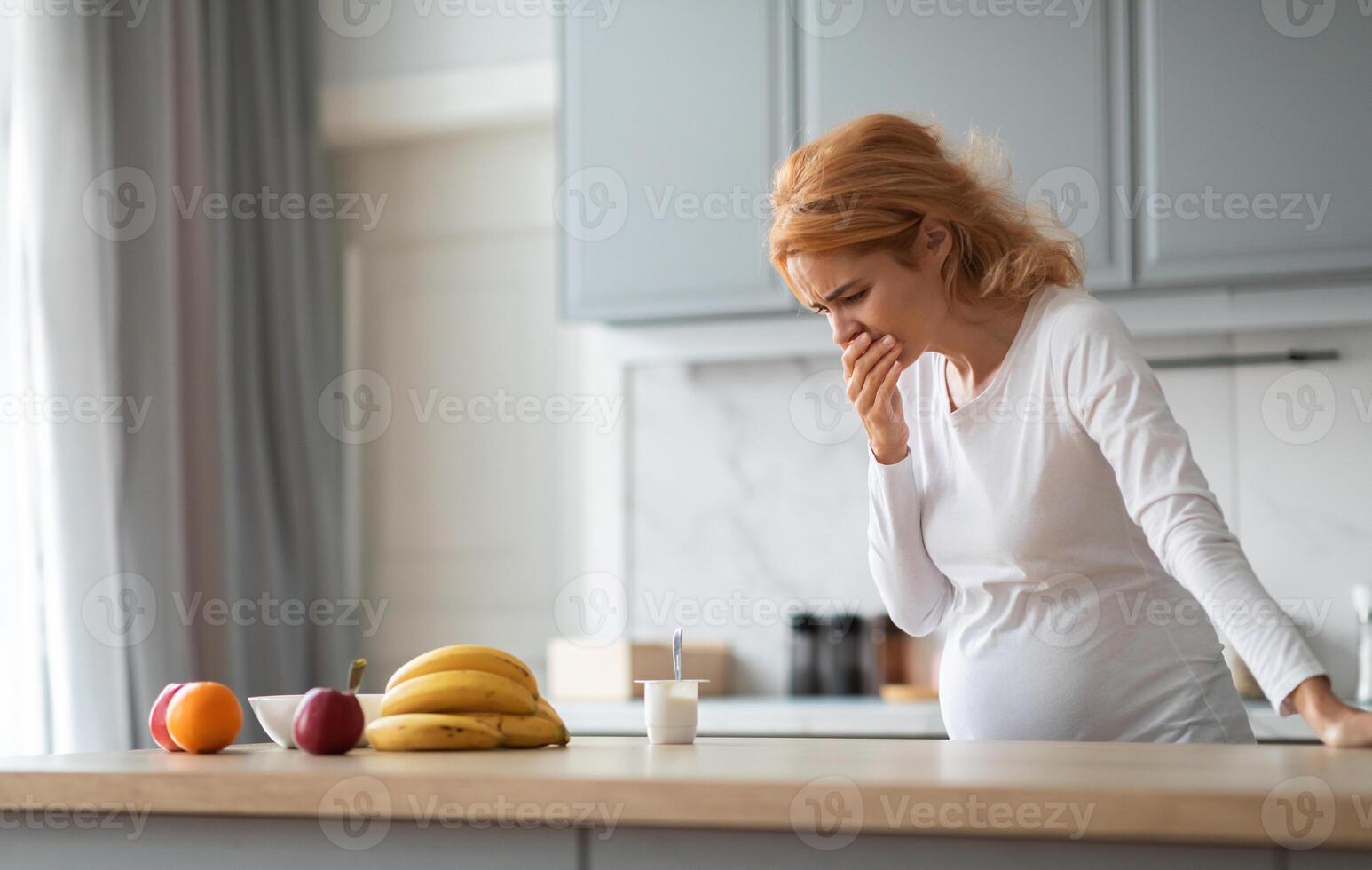 A pregnant woman stands in a stylish kitchen, visibly uncomfortable as she doubles over in nausea. Fresh fruits are arranged on the countertop, highlighting a healthy lifestyle. photo
