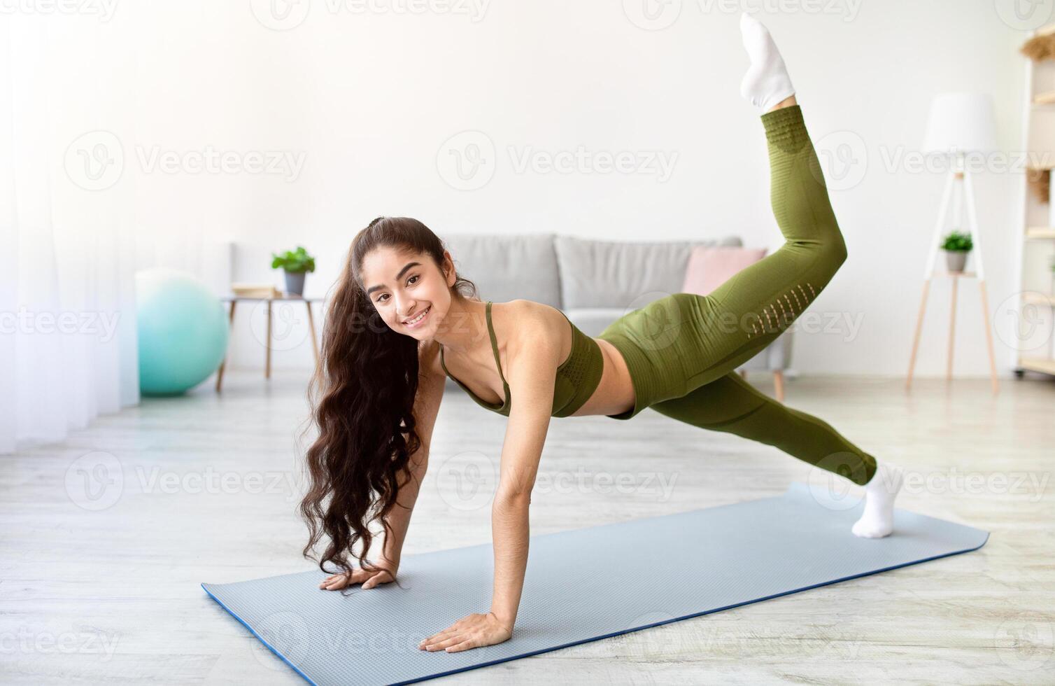 Positive young Indian woman in sportswear doing flexibility exercises, lifting her leg in plank pose at home, full length. Cool Asian lady making domestic fitness during coronavirus lockdown photo