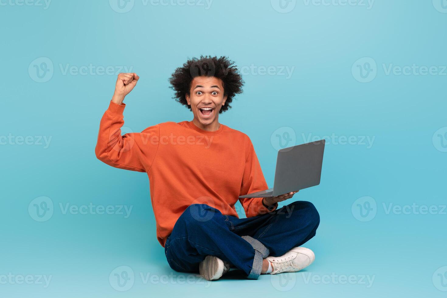 A young African American man with curly hair sits cross-legged on the floor, smiling broadly and raising his fist in triumph while looking at his laptop. The background is a vibrant blue. photo