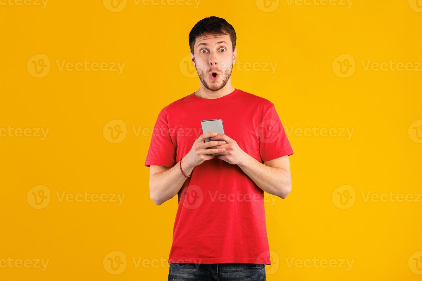 Wow, Amazing Application. Portrait of excited surprised young man using cellphone standing isolated over blue studio background, copy space. Socked guy chatting online, looking at camera photo