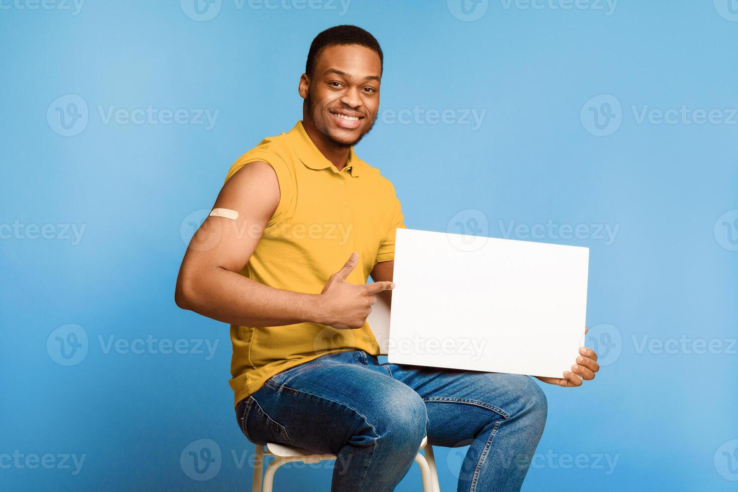 Get Your Covid-19 Vaccine. Vaccinated African Man Holding Blank Poster For Coronavirus Vaccination Ad Sitting Over Blue Studio Background. Corona Virus Prevention Concept. Mockup photo