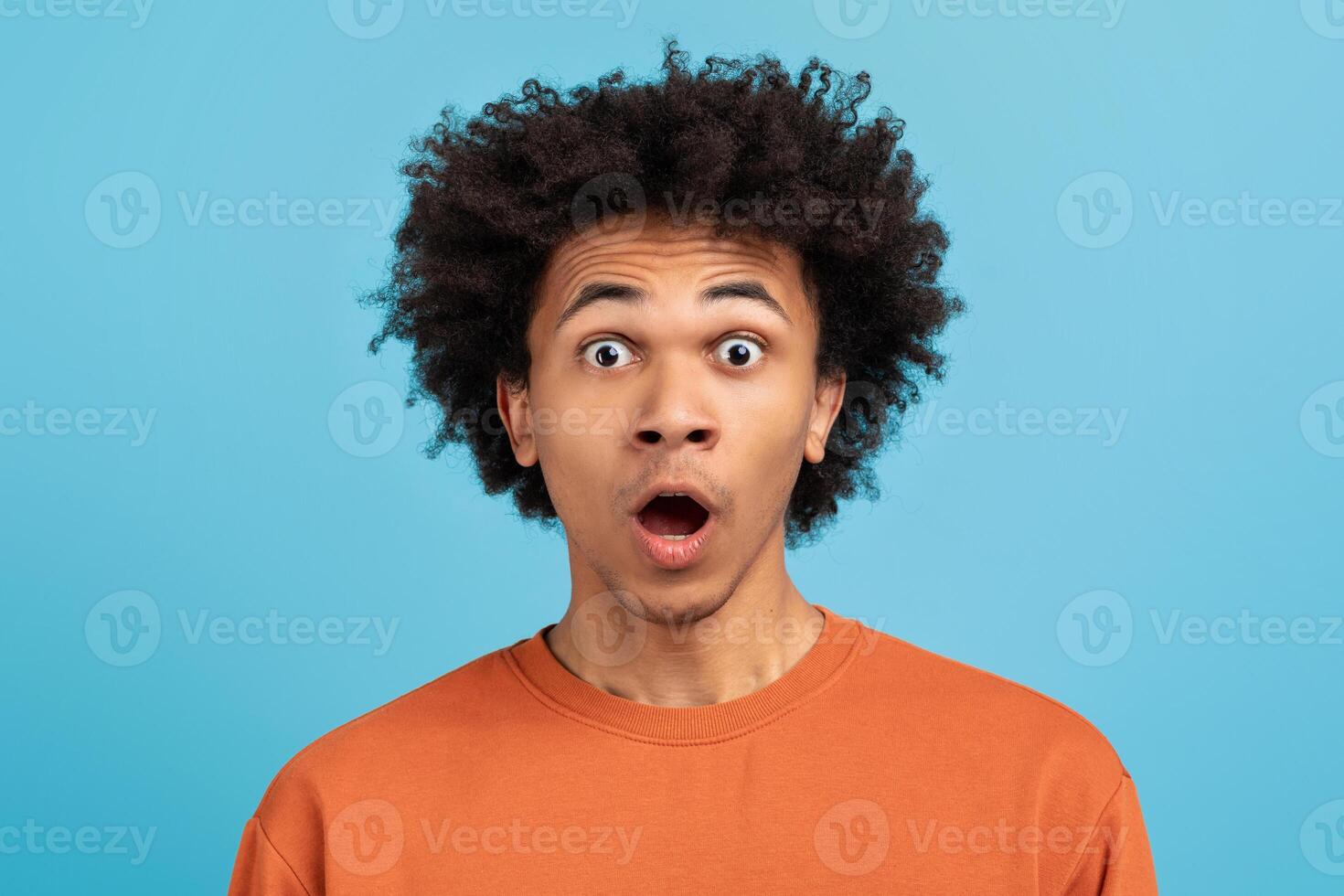 A young African American man with curly hair shows a look of surprise against a vibrant blue background. His expression conveys a strong emotion, highlighting a moment of realization or shock. photo