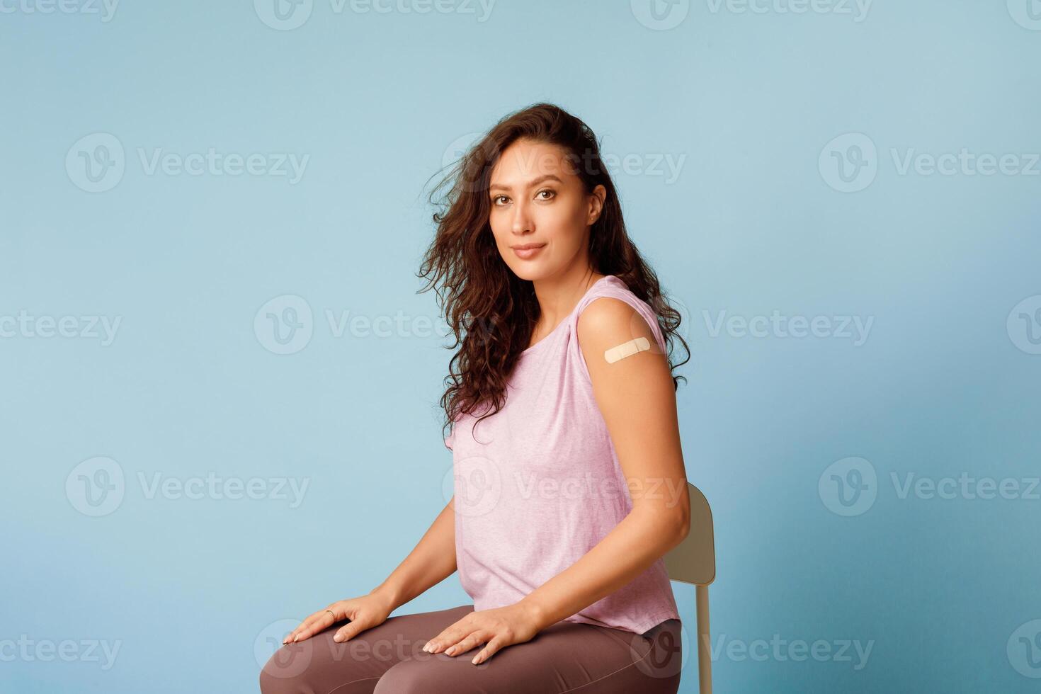 Stop Coronavirus, Vaccination Campaign. Vaccinated Woman Showing Arm Sitting With Rolled Up Sleeves Posing After Covid-19 Vaccine Injection On Blue Background. Studio Shot photo