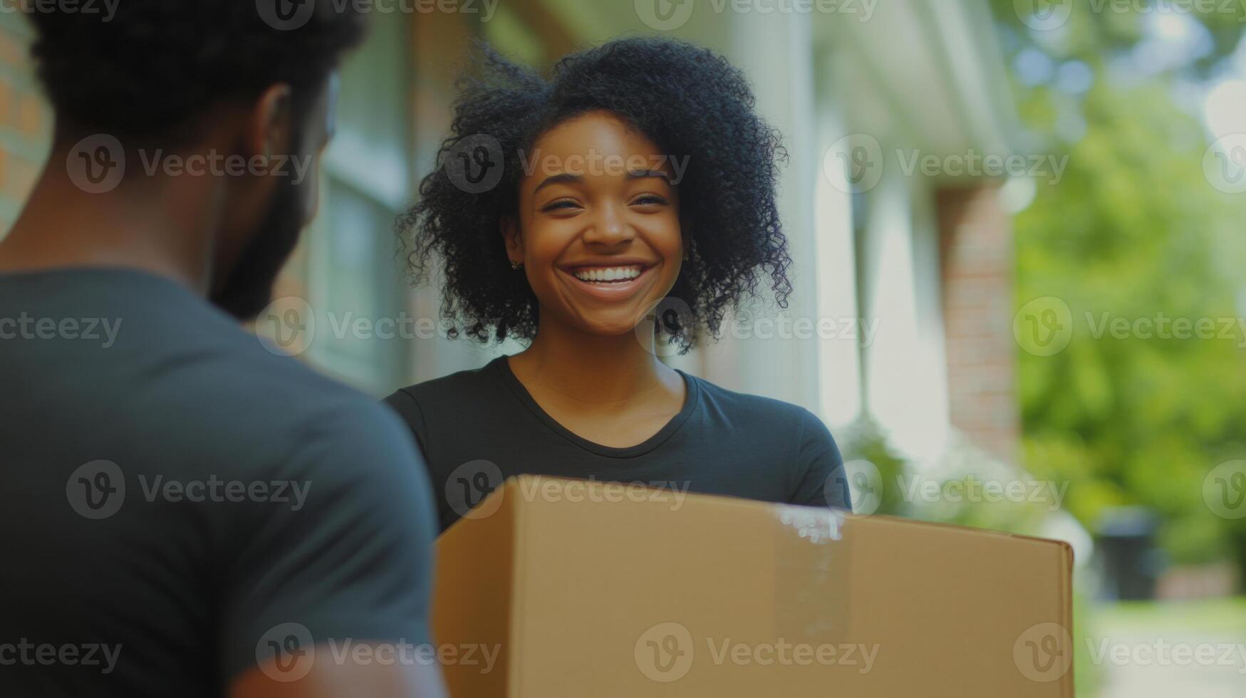 A cheerful woman greets a delivery person at her doorstep on a bright day. She happily accepts a cardboard box while standing in a well-kept residential area. photo