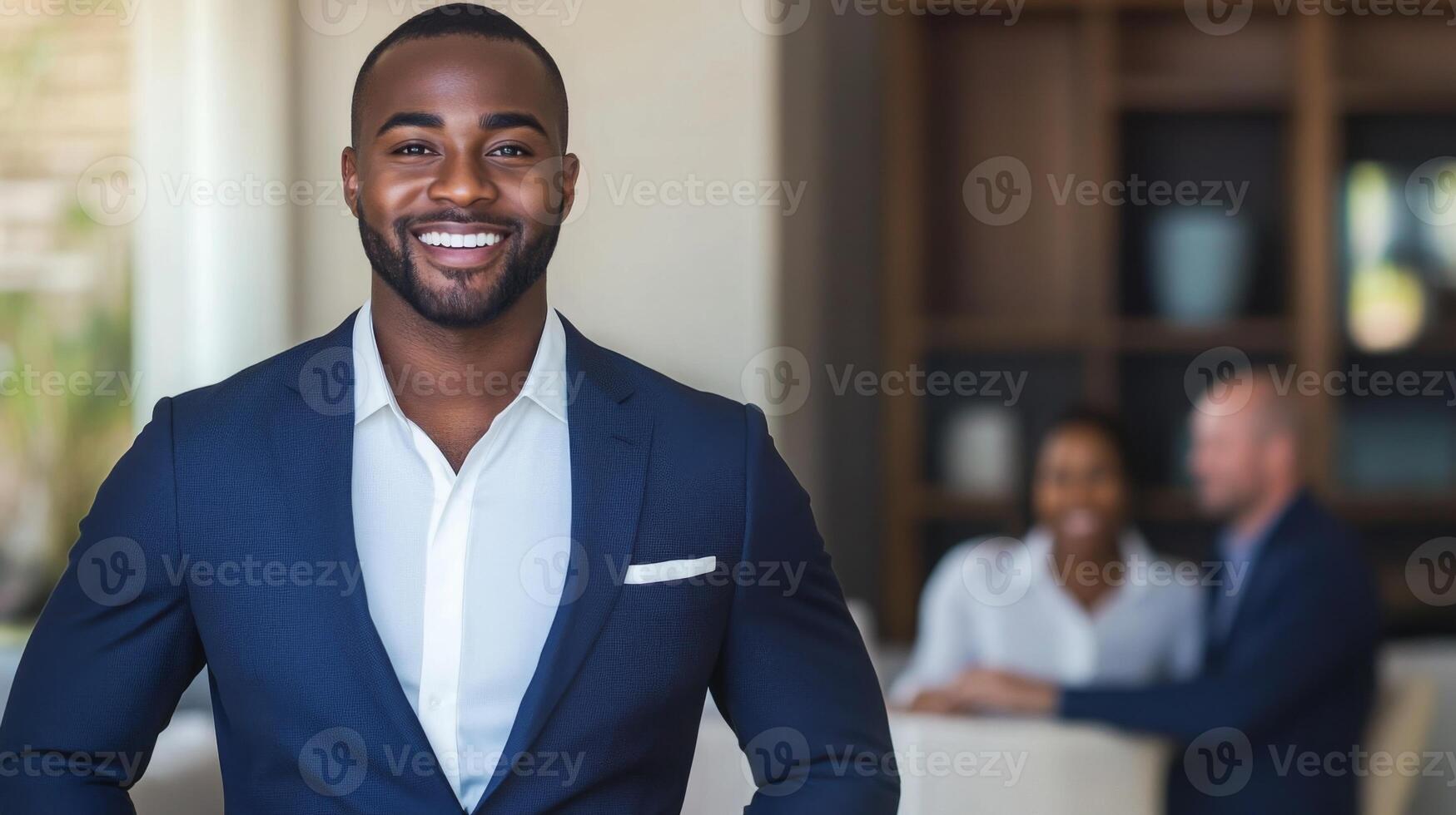 A confident real estate agent smiles in formal attire while standing in a contemporary office. Two clients can be seen engaging in conversation at a nearby table, discussing property options. photo