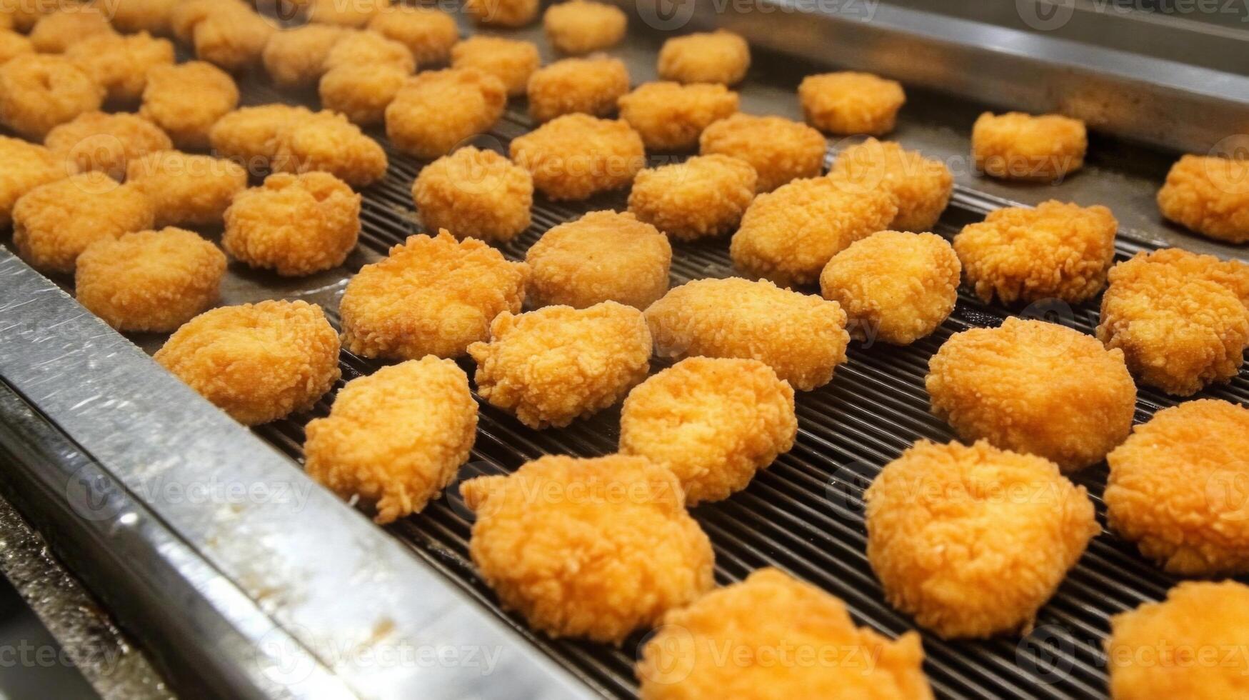 A busy food production line showcases numerous golden fried nuggets being cooked evenly. Steam rises, indicating the crispy texture and preparation of these popular snacks. photo