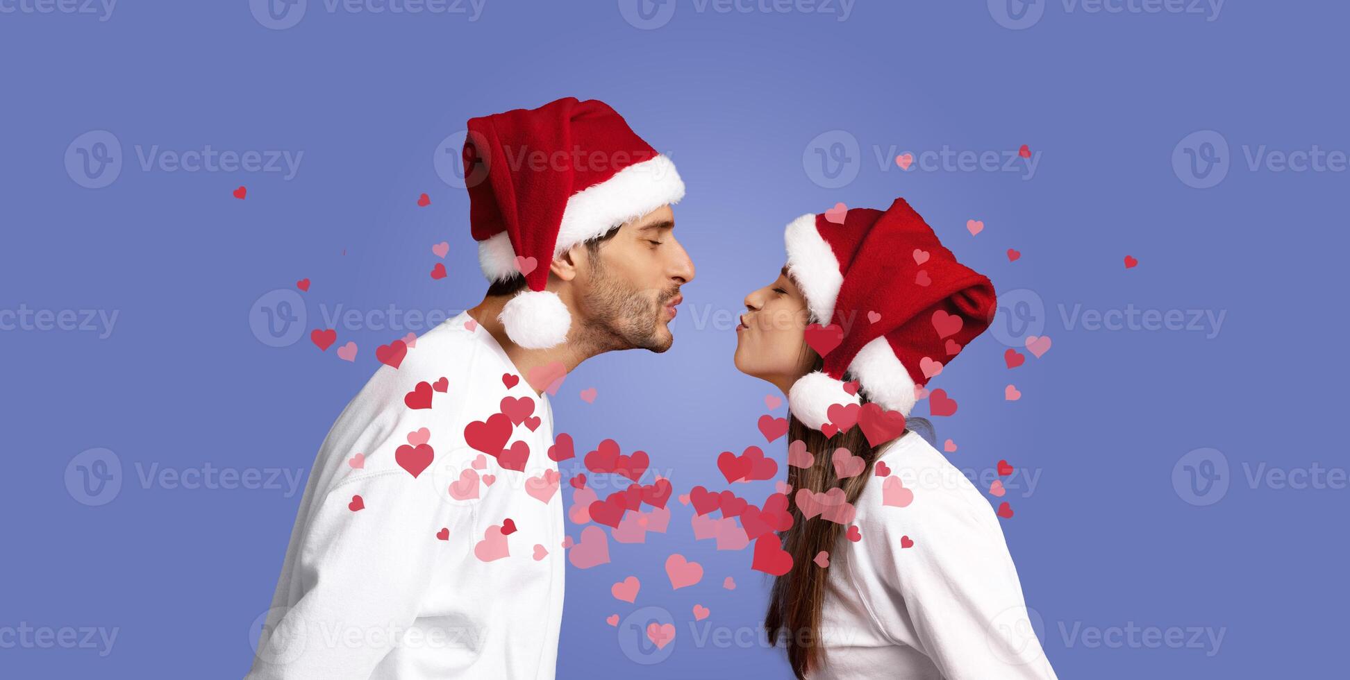 A couple in matching Santa hats leans in to share a loving kiss, surrounded by heart-shaped decorations. Their joyful expression captures the spirit of celebration during the holiday season. photo