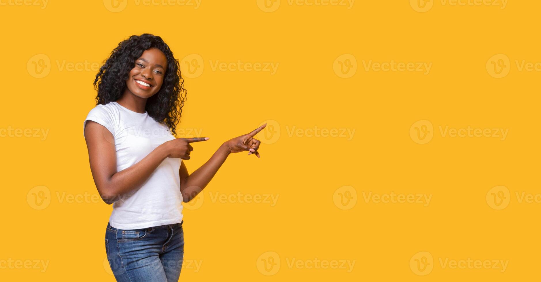 Cheerful black lady in white t-shirt pointing at copy space on yellow studio background. Smiling african american young woman showing exciting advertisement or announcement, panorama with empty space photo