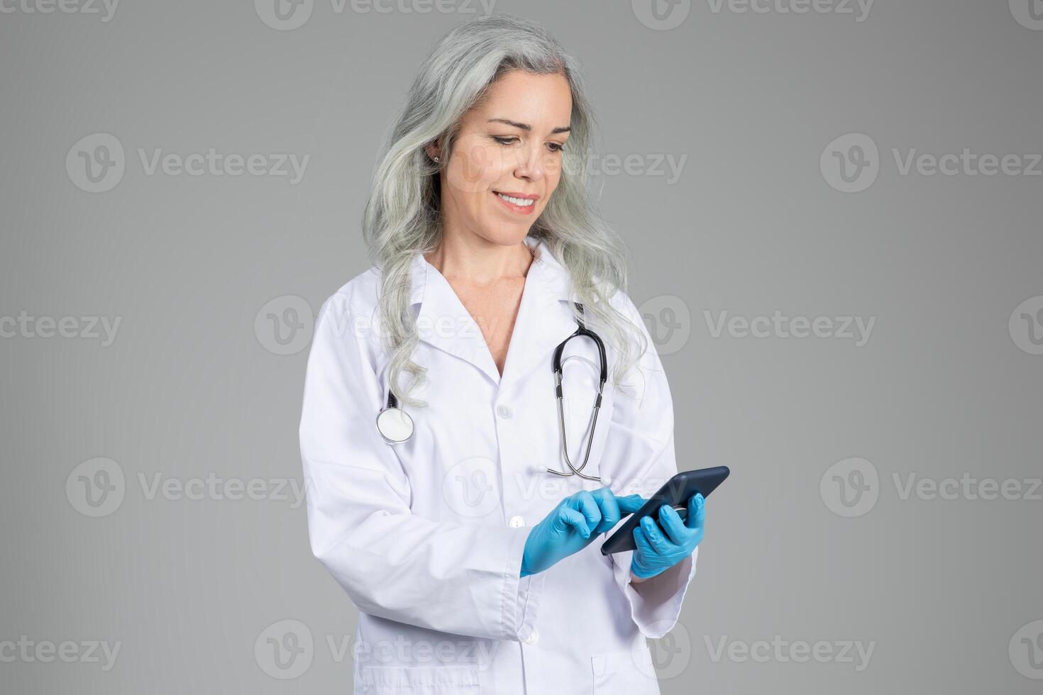 A middle aged woman in a white lab coat holds a smartphone while wearing blue gloves. She appears to be engaged in healthcare tasks. The background is neutral and unobtrusive. photo