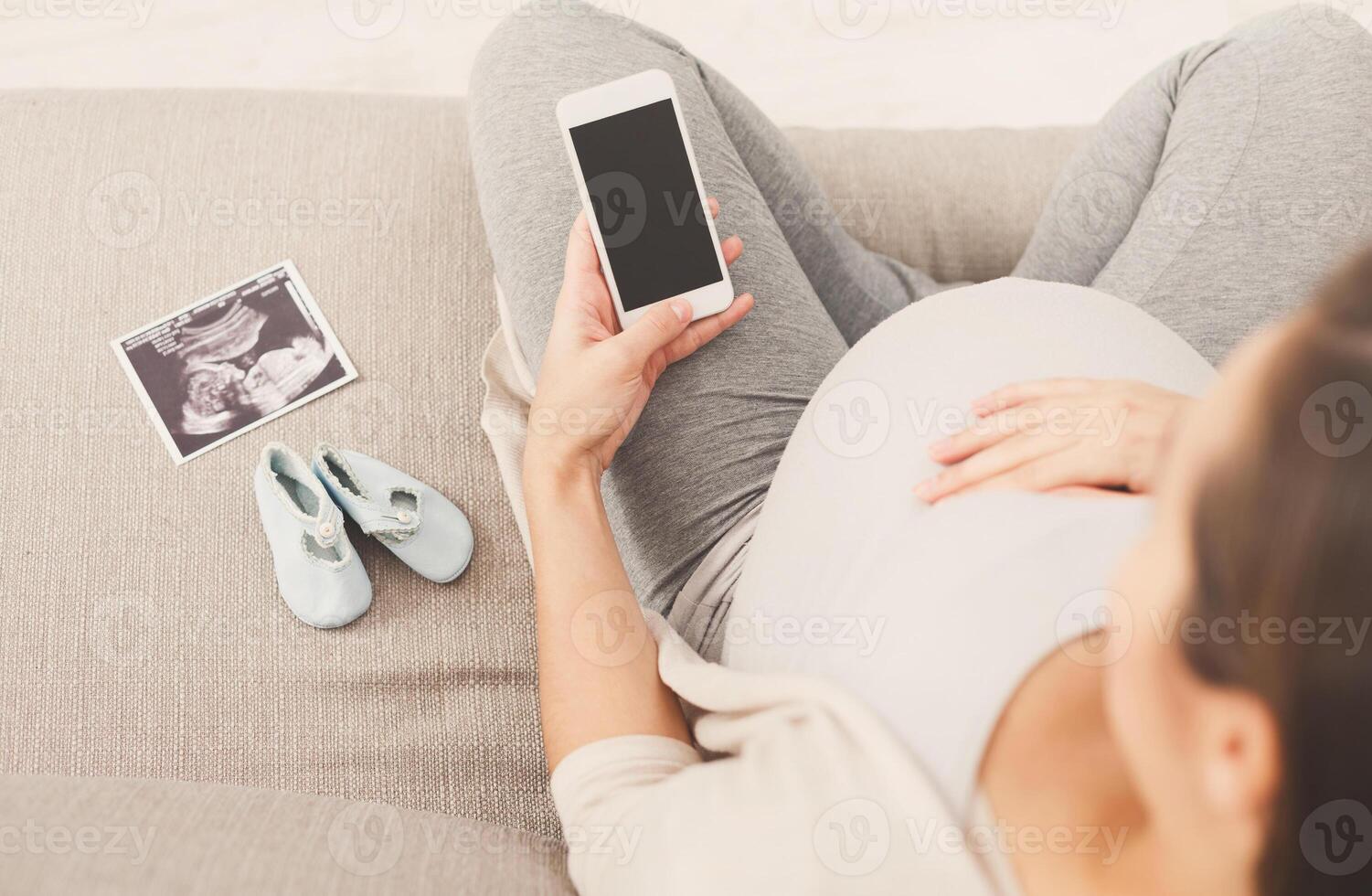 Unrecognizable pregnant woman using smartphone with blank screen, with tiny shoes and sonogram. Young expectant lady surfing net, reading about baby care, buying goods for newborn, copy space photo